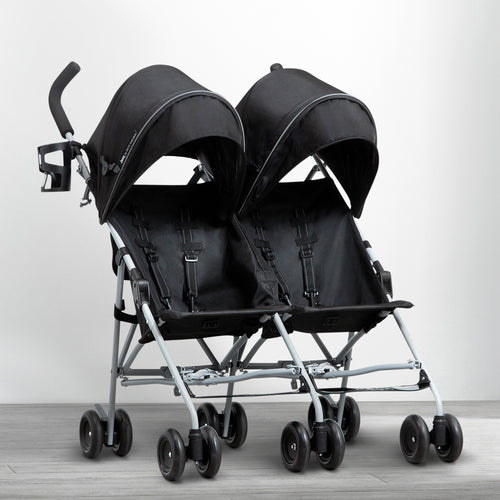A woman pushing a side by side double stroller with two children sitting in it, walking on a pathway in a park.