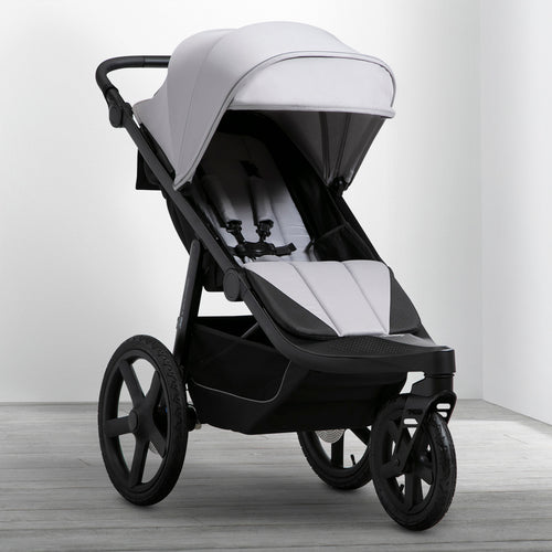 A man is pushing a black Jeep PowerGlyde Pro Jogging Stroller with a child reclined in it, walking on a trail in a forest.
