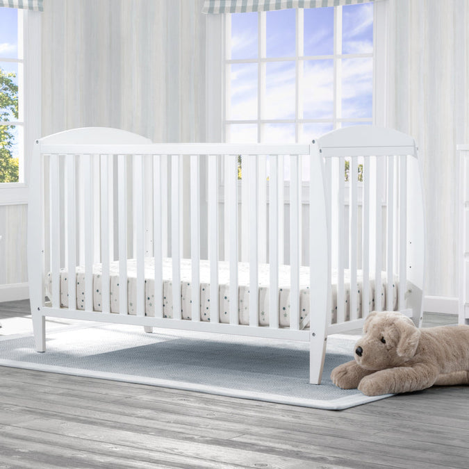 White crib with a teddy bear on a wooden floor in a bright room with large windows. 16-Taylor 4-in-1 Convertible Crib