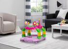 A pink and green baby activity walker toy with colorful flower and bee toys, shown in a modern living room setting. 17-Lil’ Play Station 4-in-1 Activity Walker
