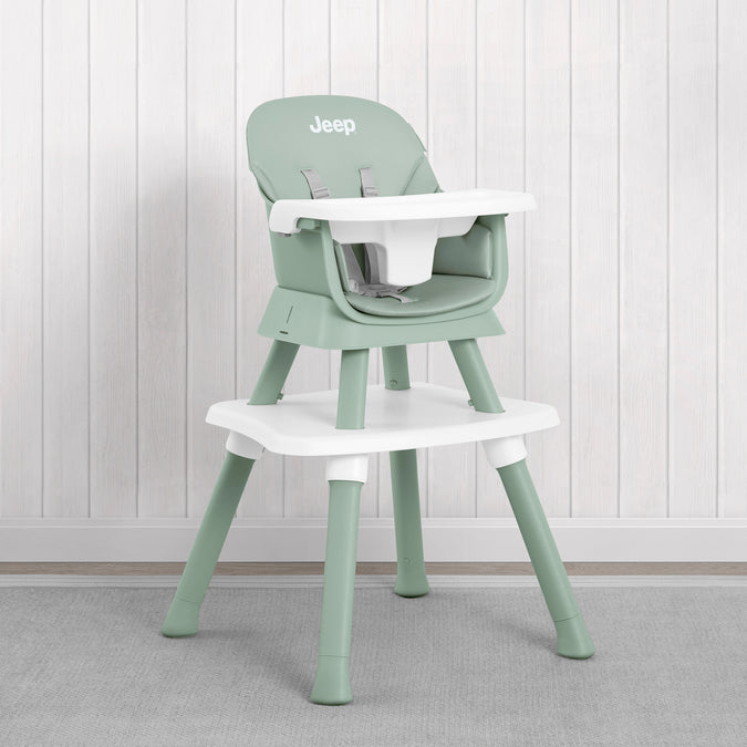 A light sage green and white plastic high chair with a matching footrest, shown against a white wood-paneled wall and gray carpet. The brand name 'Jeep' is printed on the backrest. 19-Milestone 6-in-1 High Chair
