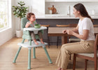 A baby sits in a light green and white high chair facing a woman sitting at a wooden dining table in a bright kitchen. 2-Milestone 6-in-1 High Chair