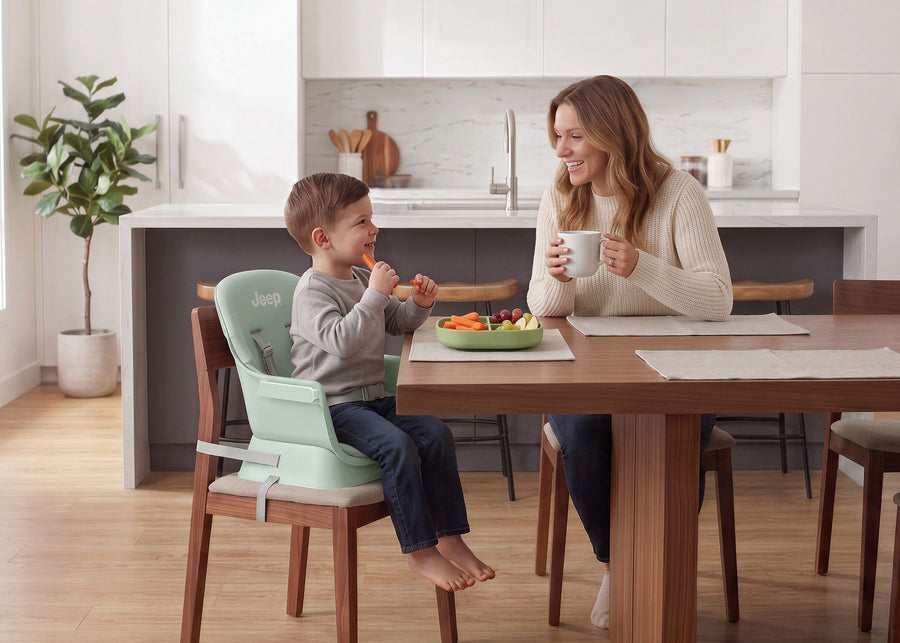 A woman and a young boy share a meal at a wooden dining table in a bright, modern kitchen. The boy is seated in a light mint green Jero toddler booster seat attached to a wooden chair, while the woman holds a white mug. 3-Milestone 6-in-1 High Chair