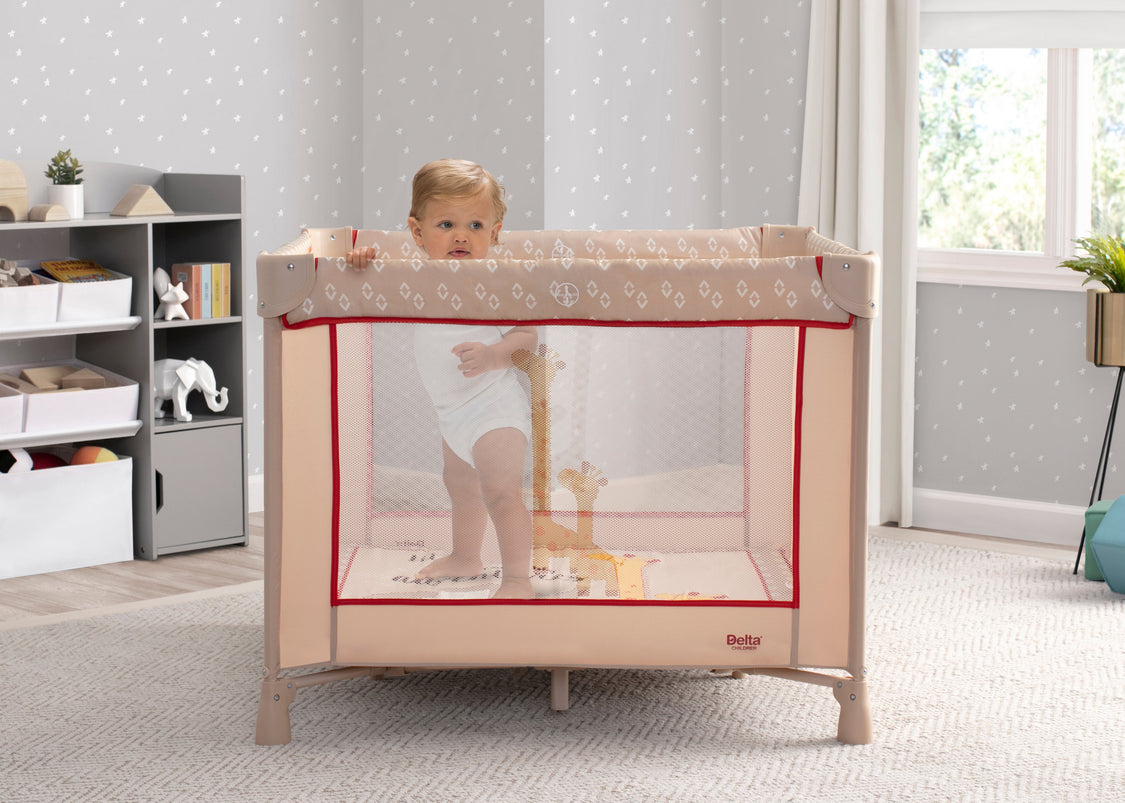 Child standing inside a portable playpen in a room with a bookshelf and window.