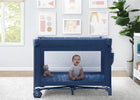 Baby sitting inside a blue crib in a room with a teddy bear and framed pictures on the wall. 7-babyGap Deluxe Play Yard