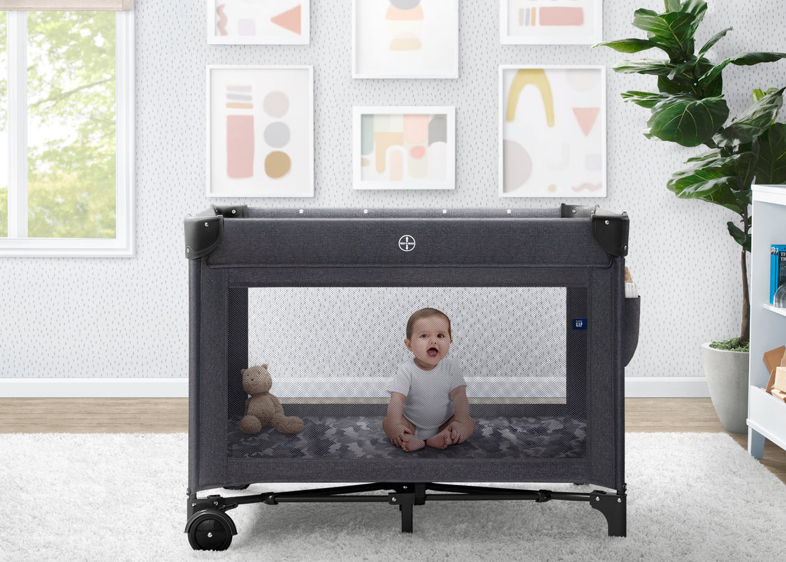 Baby sitting inside a portable crib in a room with framed art on the wall.