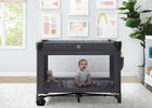 Baby sitting inside a portable crib in a room with a window, plant, and framed pictures on the wall. 14-babyGap Deluxe Play Yard