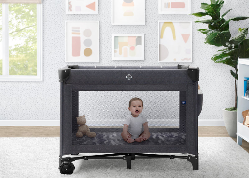 Baby sitting inside a portable crib in a room with a window, plant, and framed pictures on the wall. 14-babyGap Deluxe Play Yard