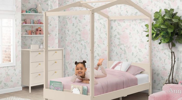Child in a house-shaped bed with pink bedding in a room with floral wallpaper and a dresser. 3-Poppy Collection