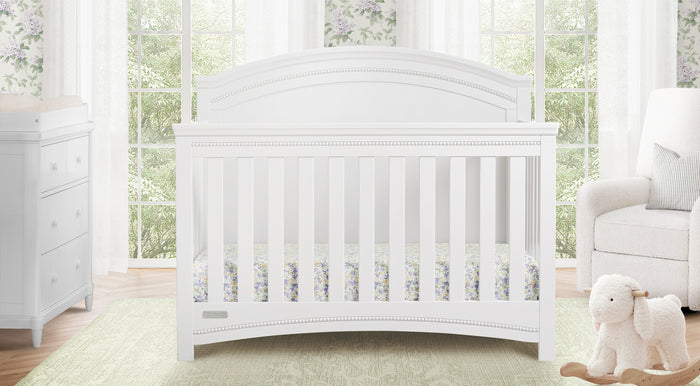 A white full-size crib with a decorative headboard, placed in a nursery room with a dresser and a glider in the background, and a mattress pad on the crib. 0-Emma Collection