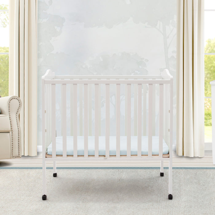 White crib in a bright nursery with large windows and curtains. 1-Folding Portable Mini Baby Crib with 1.5-inch Mattress