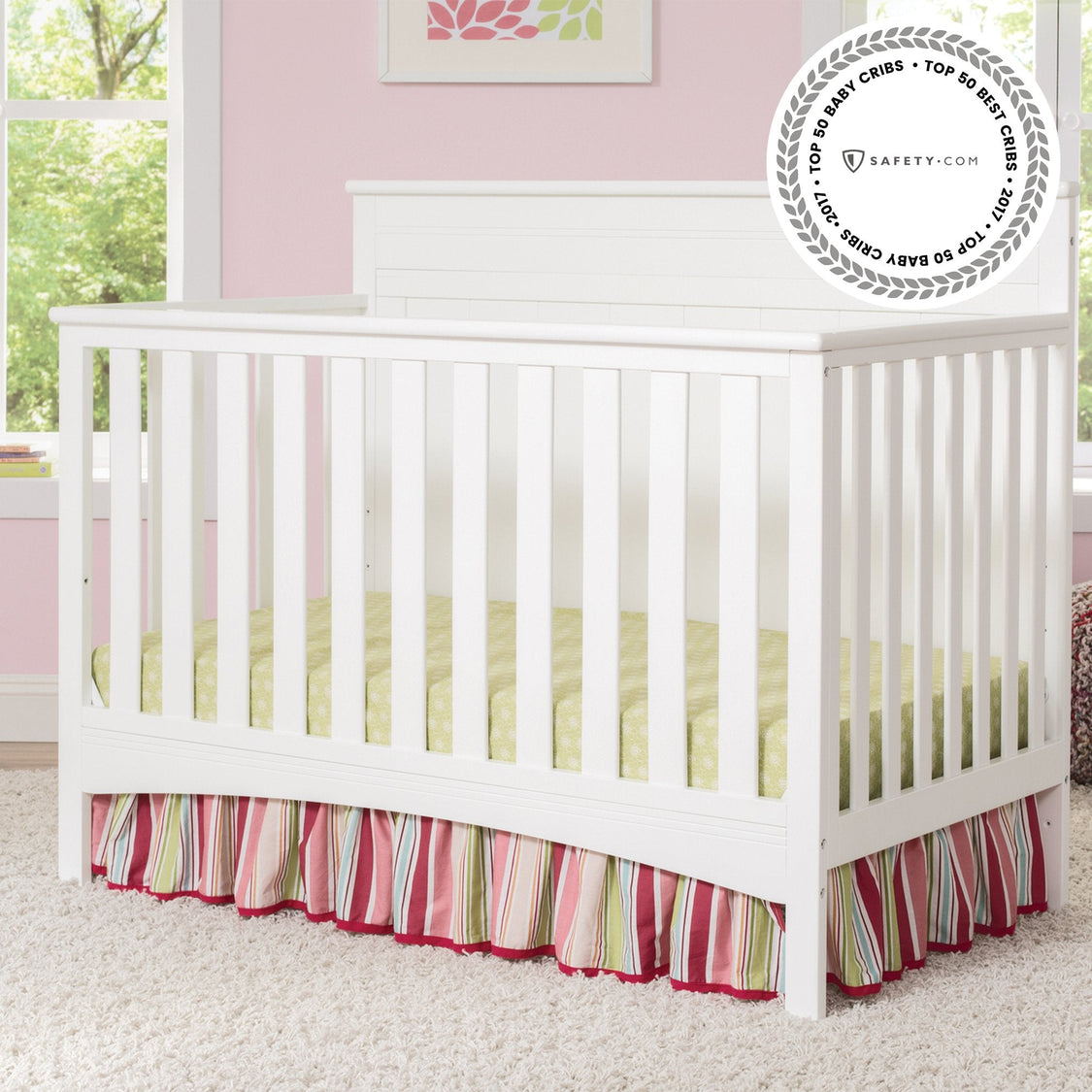 White crib with colorful skirt in a nursery with pink walls and window.