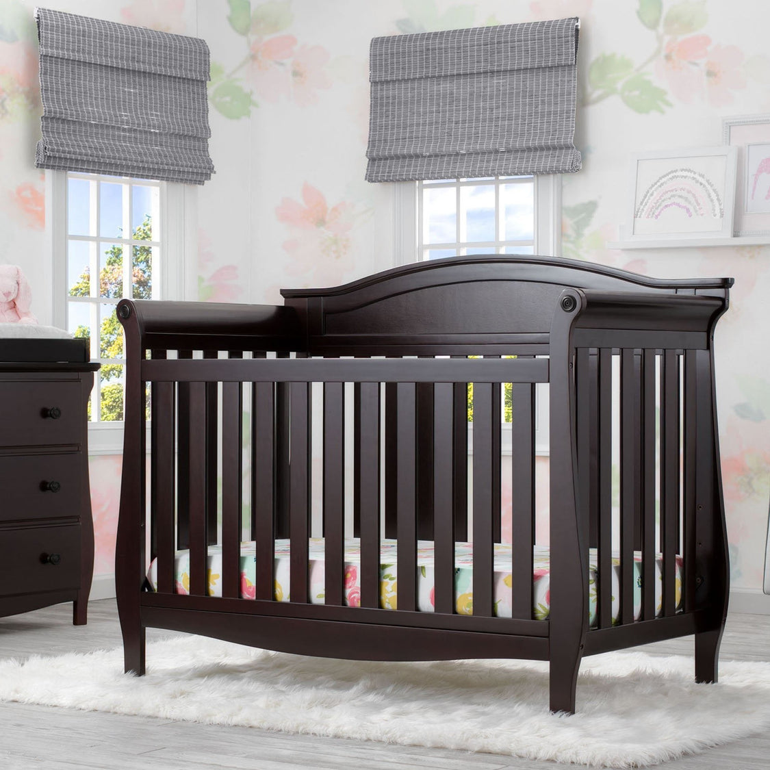 Dark wood crib in a nursery with floral wallpaper and window blinds.
