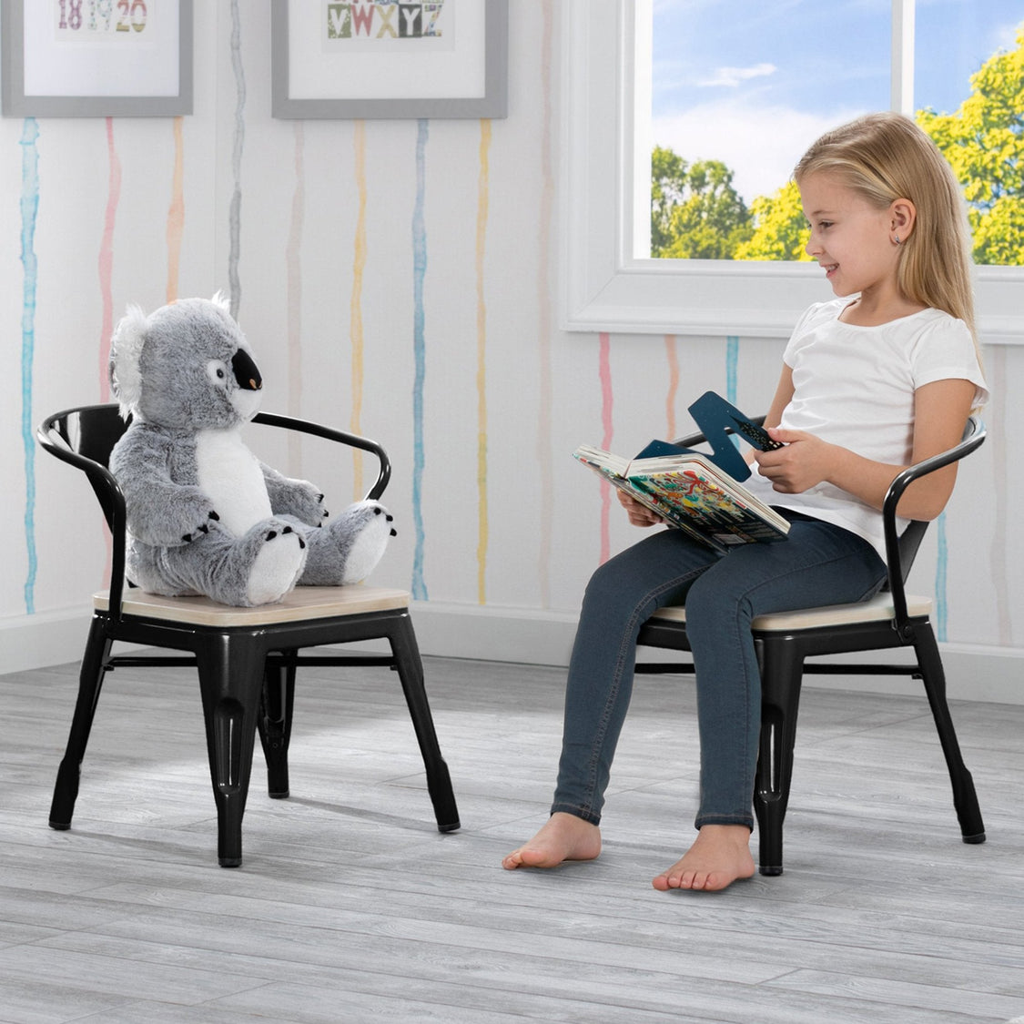 Child reading a book with a stuffed koala toy on a chair in a room with striped wallpaper and a window.