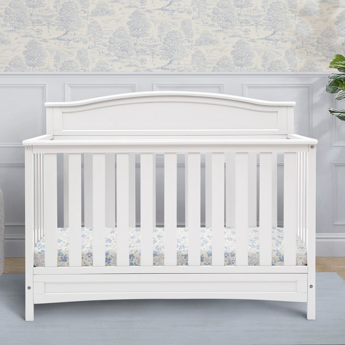 A white Emery 4-in-1 Convertible Crib with a curved headboard and airy slats, placed in a nursery room. 1-Emery 4-in-1 Convertible Crib