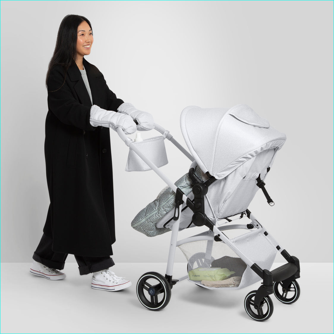A person wearing a black coat and white babyGap stroller handmuffs is pushing a white baby stroller with a bassinet. The person appears to be smiling.