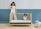 Woman standing next to a wooden crib with a child inside, in a room with blue walls and a white rug. 15-Serta CloudCool Crib and Toddler Mattress