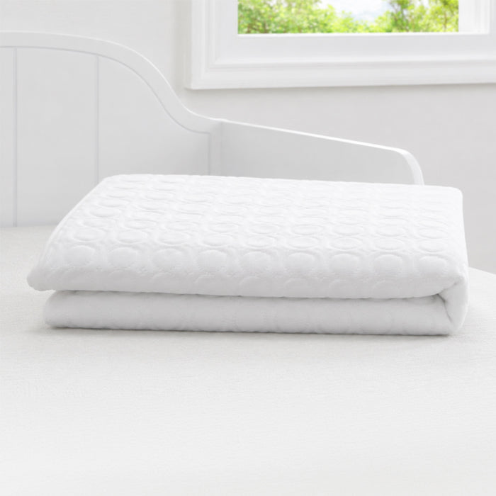 A folded white quilted mattress topper or pillowcase sits on a white bed in a bright, minimalist bedroom with a window and white headboard. 1-Eco-Cool Cooling Crib Mattress Protector