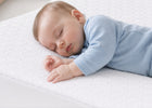 A sleeping baby wearing a light blue long-sleeve onesie, lying on a white textured mattress. 14-Eco-Cool Cooling Crib Mattress Protector