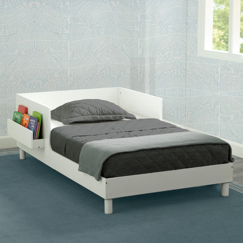 A white toddler bed with a changeable shelf on one side, placed in a room with a light blue wall and a framed picture on the wall above the bed. Next to the bed is a small chair.