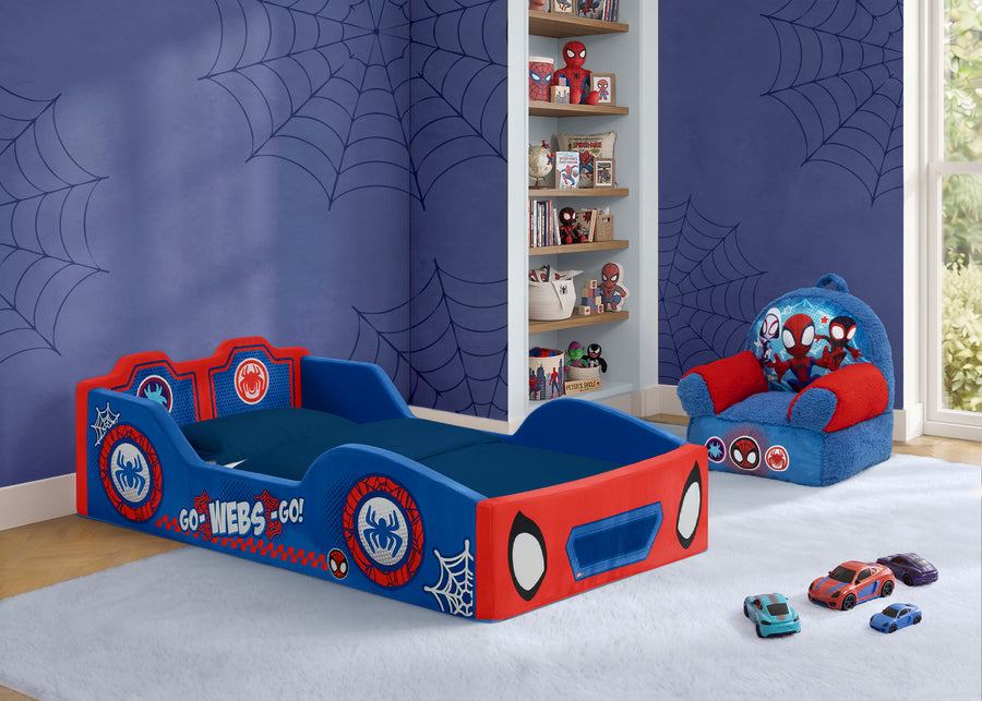 A children's bed styled with Spider-Man graphics, featuring a blue and red frame with spider web patterns and character decals. The bed is shown in a decorated bedroom with Spider-Man themed wallpaper, a matching armchair, and toy cars on the floor. 14-Spidey and His Amazing Friends Foam Frame Race Car Toddler Bed