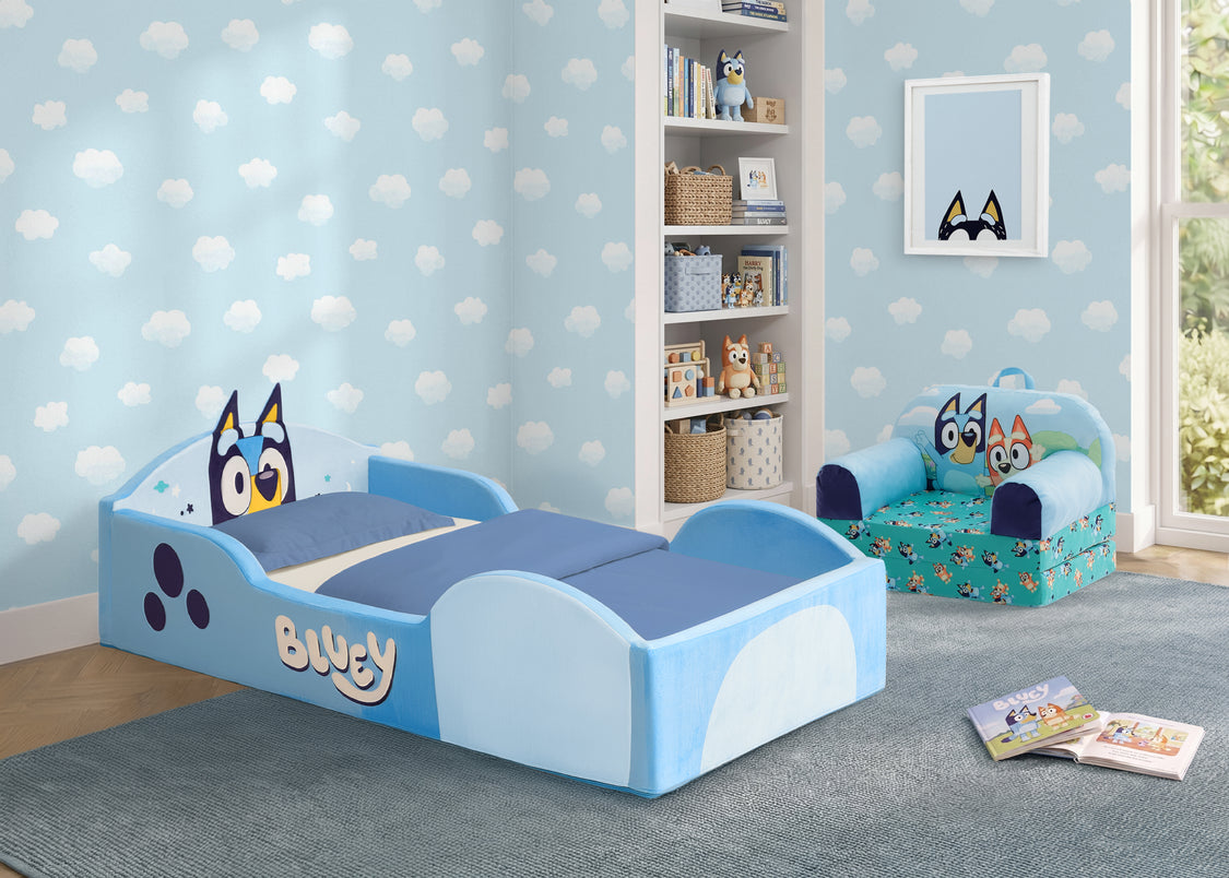 A children's bedroom scene featuring a light blue and blue toddler bed with an owl character on the headboard, a matching patterned armchair, light blue cloud-patterned wallpaper, and a white shelving unit in the background.