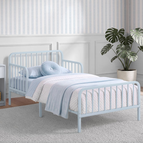 A light blue metal toddler bed with a guard rail, styled with a light blue and white patterned duvet, matching pillow, and a round tufted pillow, set in a child's bedroom with striped wallpaper and a potted plant.