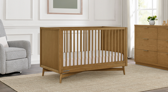 A white full-size crib from the Penelope Collection placed in a nursery room with a dresser and glider in the background. 2-Dylan Collection