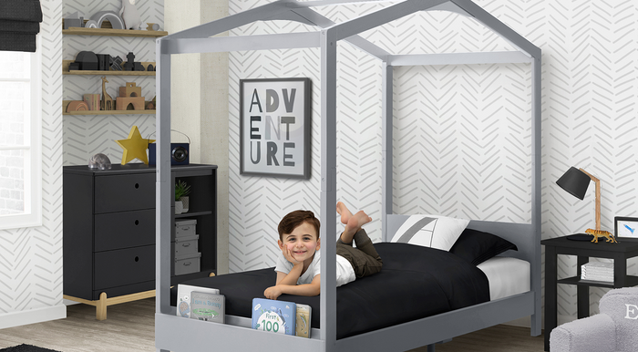 A child lying on a gray canopy bed frame in a room with furniture and decorative wall stickers. 0-Poppy Collection