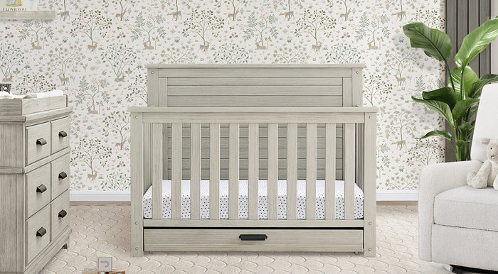 Gray crib with white mattress in a nursery setting with wallpaper and furniture. 1-Caden Collection