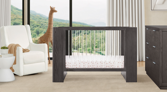 A modern white nursery set including a crib with a white guardrail, a white glider chair, a dresser, and a changing top, set in a room with a large window and a stuffed animal on the chair. 2-Aerin Collection