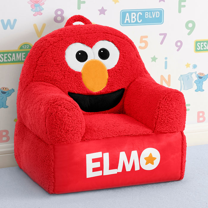 A plush red children's armchair designed like Elmo from Sesame Street, with white eyes, a yellow-orange nose, a black mouth, and the name 'ELMO' printed on the front. The chair is photographed in a child's room setting against a decorated wall. 3-Sesame Street Elmo Cozee Buddy Chair