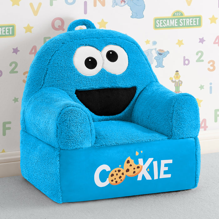 A plush blue children's armchair designed like the Cookie Monster from Sesame Street, with large white eyes, a black mouth, and 'COOKIE' printed on the front with cookie graphics. The chair is photographed in a styled room setting with a patterned wall behind it. 0-Sesame Street Cookie Monster Cozee Buddy Chair
