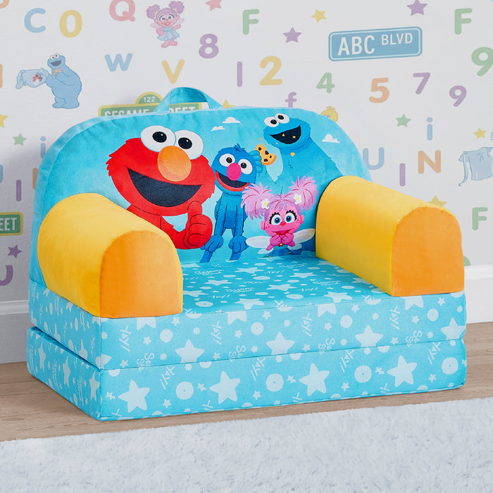 A children's fold-down play couch or small bed in a Sesame Street theme, featuring Elmo, Grover, Abby, and Cookie on a light blue star-patterned seat, with yellow armrests, set against a child's room wall with alphabet letters. 0-Sesame Street Cozee Buddy Flip-Out Chair