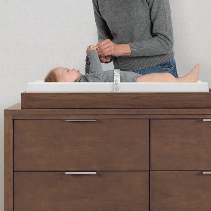 A baby changing table in dark brown wood with a white changing pad, shown with a baby and an adult standing behind it against a light gray wall. 9-EasyChange Changing Pad