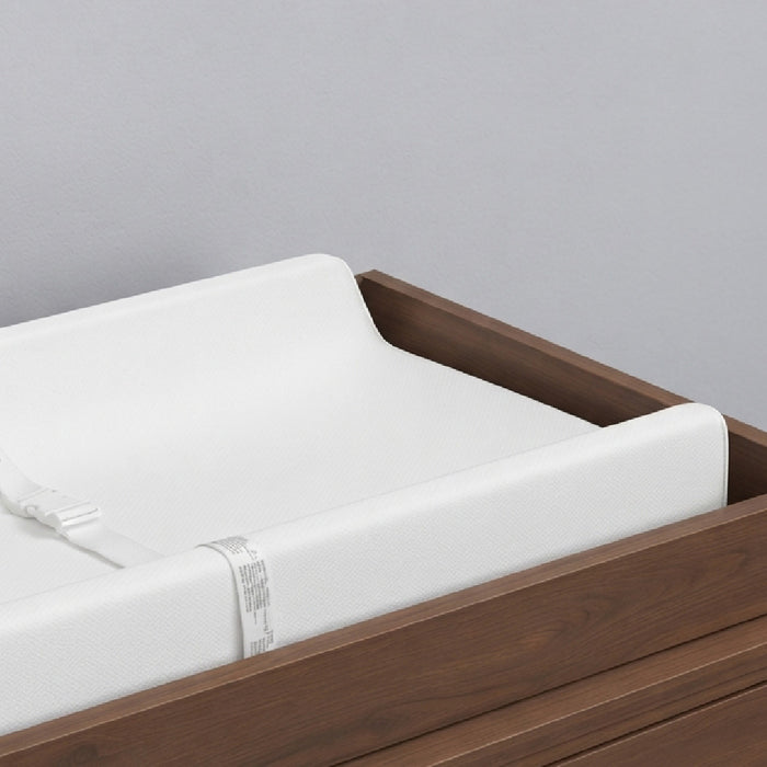 A close-up of a white changing pad with a strap, set inside a dark brown wooden changing table against a light gray background. 3-EasyChange Changing Pad