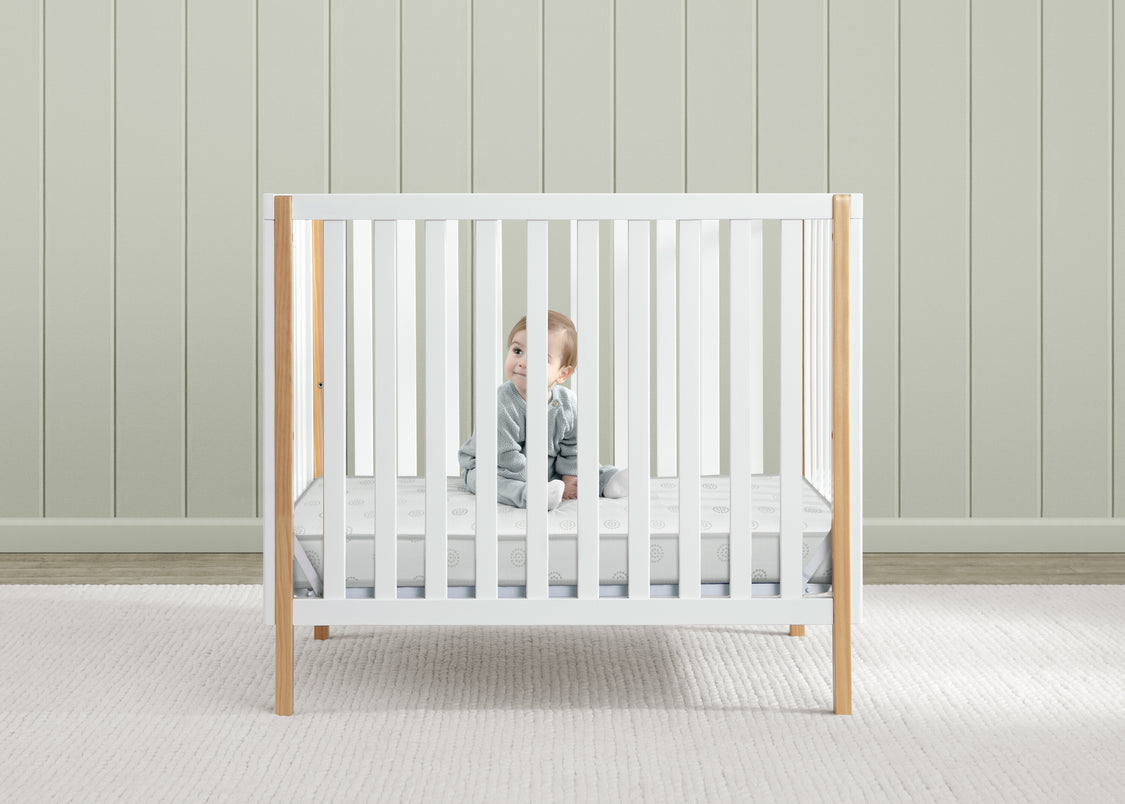 Child in a white crib with wooden frame against a light wooden paneled wall.