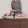 A baby in a gray outfit lies on a beige curved changing pad placed on a dark wood dresser, while an adult in a gray sweater adjusts the baby's clothing. The scene is indoors against a light textured wall. 10-Beautyrest Molded Waterproof Changing Pad with Fresh Technology