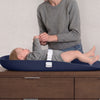A baby in a gray outfit lies on a navy changing pad on a wooden dresser while an adult in a gray sweater adjusts the baby's harness. The background is a light beige textured wall. 10-Serta Molded Waterproof Changing Pad with Fresh Technology