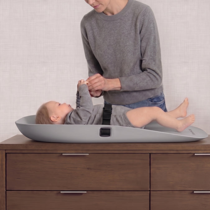 A baby in a gray outfit lies on a gray changing pad placed on a wooden dresser, while an adult in a gray sweater adjusts the baby's clothing. The scene is indoors against a light textured wall. 10-Molded Waterproof Changing Pad with Fresh Technology
