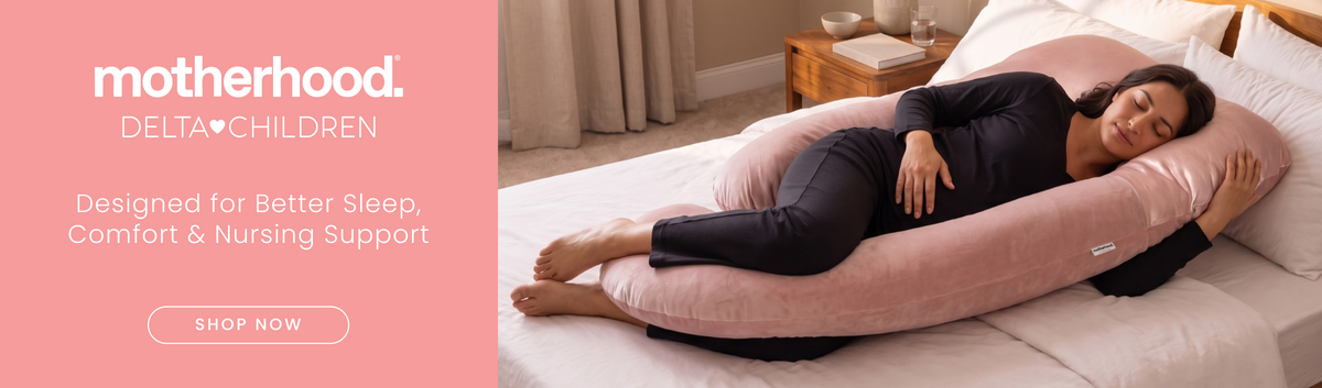 A woman lies on a bed using a large pink U-shaped nursing pillow for support. The left side has a pink banner with the text 'motherhood.