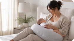 A woman in a beige nursing gown and pants holds a baby on a white nursing pillow while breastfeeding in a softly lit bedroom.