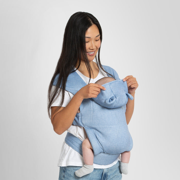 A woman is seen from the back, wearing a pink baby carrier with a sleeping hood. The carrier is designed with a supportive waistbelt and spreadable cross straps, and it features a detachable sleeping hood with Brannan Bear-inspired details. 0-babyGap Legacy Baby Carrier