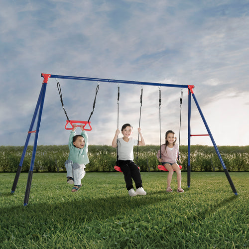 Two children are swinging on red swings attached to a blue metal A-frame swing set with a trapeze bar in a grassy outdoor area.