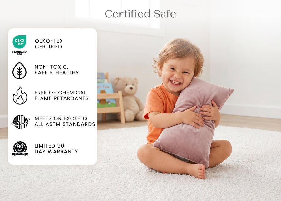 A toddler in an orange shirt sits on a white carpet in a bright room, hugging a dusty pink pillow. A large text panel on the left lists product certifications including OEKO-TEX Certified, Non-Toxic, Safe & Healthy, Free of Chemical Flame Retardants, Meets or Exceeds All ASTM Standards, and Limited 90 Day Warranty. 21-Plush Comfort Toddler Pillow