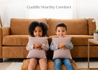 Two children sitting on a brown sofa in front of a matching cushion, each holding a light pink pillow. The text 'Cuddle Worthy Comfort' is displayed at the top of the image. 24-Plush Comfort Toddler Pillow