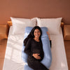 A pregnant woman lies on a light blue U-shaped maternity pillow on a bed with white sheets and pillows, photographed from above against a warm brown wall. 46-U-Shaped Pregnancy Pillow with Customizable Head Pillow