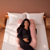 A pregnant woman lies on a bed using a large pink crescent-shaped maternity pillow, with white pillows and wooden bedside tables against a terracotta wall. 42-U-Shaped Pregnancy Pillow with Customizable Head Pillow