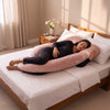 A woman lies on a large pink maternity support pillow on a bed with white linens, in a softly lit bedroom with a wooden headboard and bedside table. 40-U-Shaped Pregnancy Pillow with Customizable Head Pillow