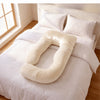 A cream-colored U-shaped nursing pillow resting on a neatly made bed with white sheets and pillows in a bright bedroom. 50-U-Shaped Pregnancy Pillow with Customizable Head Pillow